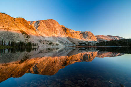 Morning sunlight illuminates the mountains and reflects off the calm waters of an alpine lake.