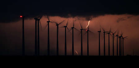 The silhouette of spinning wind turbines is backlit by a bolt of lightning in a thunderstorm at...