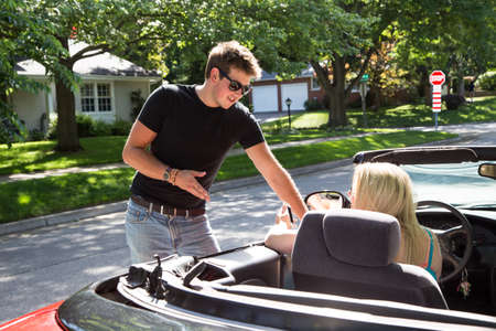 A young man stands talking to a young lady in a red convertible.  He may be giving her directions or...