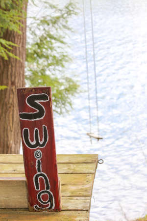 hand painted sign marks the jumping off point for the tree swing at the cottage.  Empty swing not in use hangs over the lake belowの写真素材