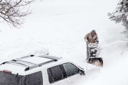 A man in his driveway clearing snow with a snowblower, taken from a snowy window aboveの写真素材
