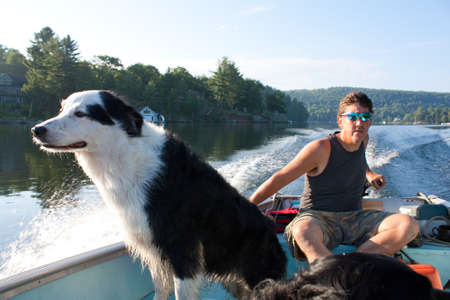 Man steers fishing boat through the waters of Lake of Bays, as his dog stands up sniffing the breezeの写真素材