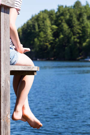 a generic young woman reads a book on the dock overlooking the lake at the cottageの写真素材