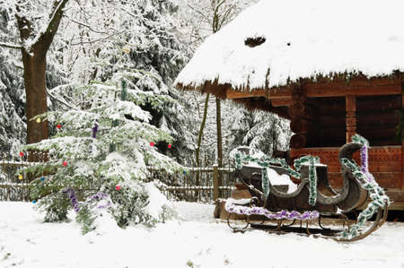 Dressed Christmas tree, old wooden cottage and Santa Claus sleigh in a tranquil winter forest. Christmas outdoor background.の写真素材