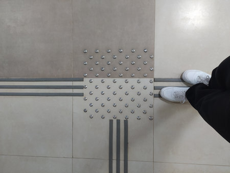 Feet in white sneakers in front of tactile tiles for visually impaired people on the floor indoors.の写真素材