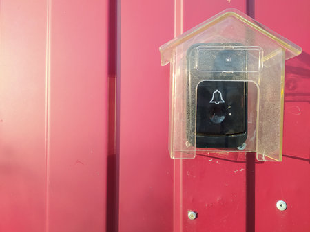Close-up of a plastic protective case over a doorbell on a red metal gate at the entrance to a...