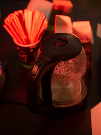 Electric glass kettle with condensation droplets on transparent body sits on dark table beside red...