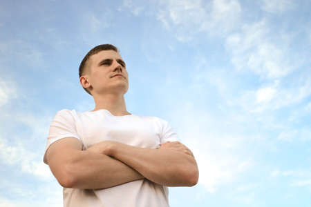 The guy in a white T-shirt is standing against the blue sky with his arms crossedの写真素材