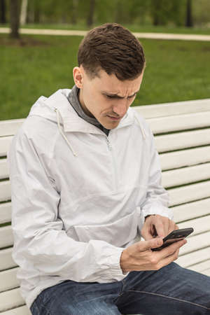 a young guy of the European appearance sits on the bench and dials a message on the phoneの写真素材