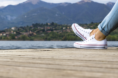 Legs and feet of a young girl on a wooden pier with white shoes in front to the lakeの写真素材