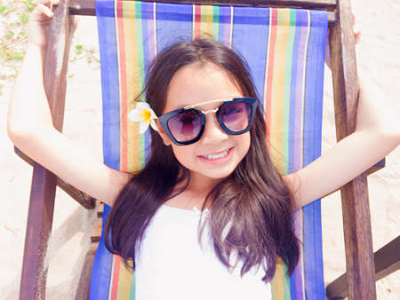 Asian long black hair girl is laying down on the colorful chair. She is smiling and wearing black sunglasses and white flower behind the right ear.の写真素材