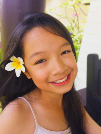 Asian long black hair girl is smiling and wearing white flower behind the right ear. She is sitting on the wooden chair.の写真素材