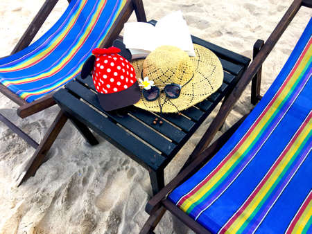 Red cap with white polka dots decorated with red bow and black fabric , straw hat with black leather rope, flower, opened book and black flame sunglasses, that located on black wooden table near the colorful canvas chair. All this item is a list of tourist attractions.の写真素材