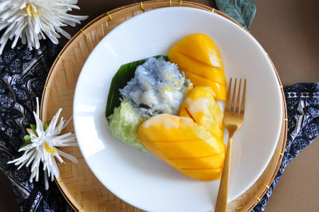 Top view of popular Thai dessert sweet Mango with sticky rice topping with coconut milk serve on white dish with golden fork put on handmade trayの写真素材
