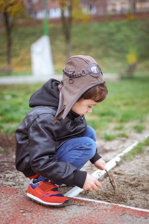 Happy boy in a pilots cap and brown jacket plays with with sand in an autumn park and dreams.の写真素材