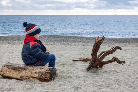 Cute boy in winter jacket and knitted hat siting from behind on deserted beach on log next to snag.の写真素材