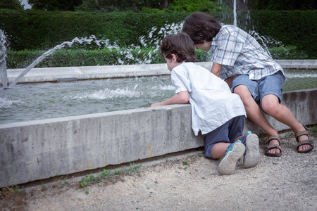 Two brothers are sitting in a summer park near a fountain and playing in the water. Family have fun.の写真素材