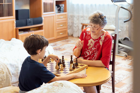 A grandmother teaches her young grandson to play chess at home. The boy listens attentively while making a move, capturing a warm and educational family interaction.の写真素材