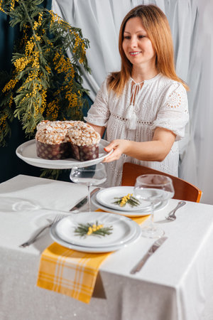 Woman in a white dress sets a festive table with Traditional Easter Colomba and mimosa. Spring mood and cozy atmosphereの写真素材