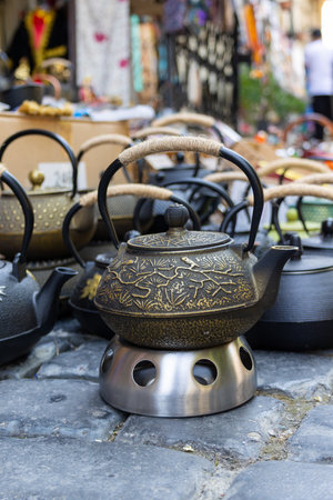 Ornate metal teapots with rope wrapped handles sit on a metal stand, creating a captivating display of traditional Asian craftsmanship in a bustling outdoor market settingの写真素材