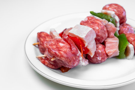 Close up raw meat and vegetable skewers featuring pork, salami, peppers, and onions on wooden sticks, arranged on a white plate against a minimalist background, ready for grillingの写真素材