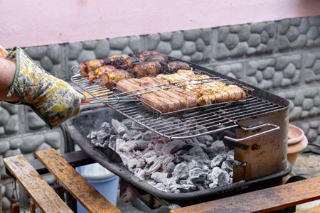 Close up hand of chef wearing a heat resistant glove skillfully adjusting meats and potatoes on a charcoal barbecue grill, creating a delicious outdoor meal for family and friendsの写真素材