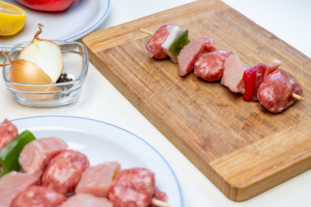 Raw meat and vegetable skewers are being prepared on a wooden cutting board. With additional ingredients like onions. Peppers. And lemon arranged nearby on white plates. Suggesting a culinary settingの写真素材