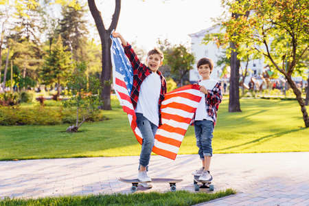Two cute brothers guy americans standing in a city park on skateboards and holding flag of USA.の写真素材