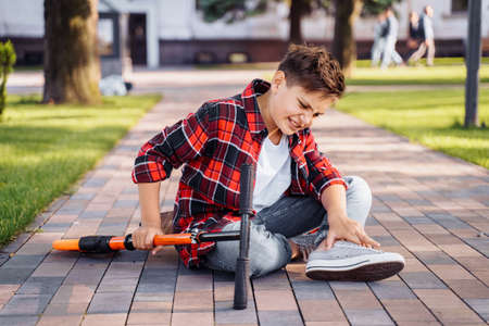 A little boy 8 years riding scooter and fell. Sitting on asphalt, holding hands on his injured leg and crying in pain. Broking his knee. It's an unexpected incident. Summer walks in the parkの写真素材