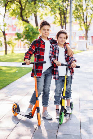 Two attractive european boys brothers, wearing red and white checkered flannek shirts, standing on scooters in the park. They laughing, smiling, hugging and having fun. Showing big fingers up sign.の写真素材