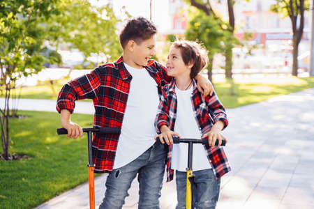 Two attractive european boys brothers, wearing red and white checkered flannek shirts, standing on scooters in the park. They laughing, smiling, hugging and having fun. Showing big fingers up sign.の写真素材