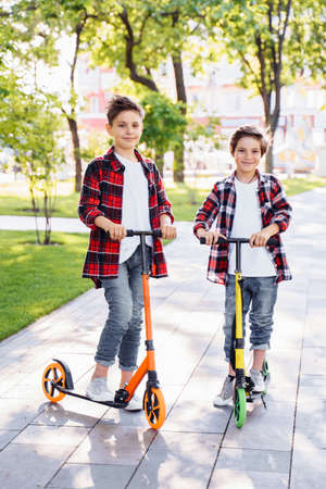 Two attractive european boys brothers, wearing red and white checkered flannek shirts, standing on scooters in the park. They laughing, smiling, hugging and having fun. Showing big fingers up sign.の写真素材