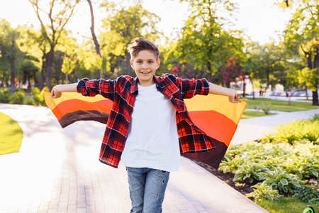 Guy of 9 years old riding skateboard on sunny road in the evening in the city park. Holding fluttering flag of Germany. Happy German day. German language, European education.の写真素材