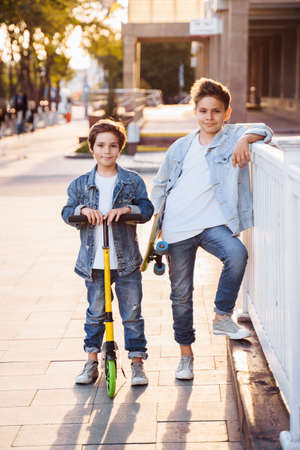 two attractive teenagers of Caucasian nationality 7 and 9 years old standing on sunny evening street in the city. Laughing, pointing fingers forward, holding scooter and skateboard.の写真素材