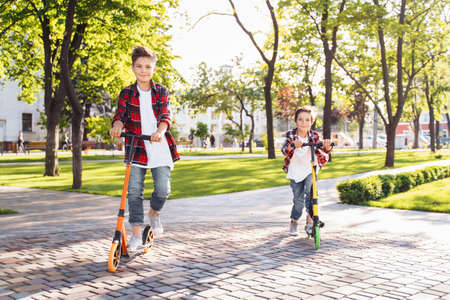 Two happy smiling teenagers brothers, 7 and 8 years old, with speed riding a scooter in the park on a summer evening. Dressed in plaid checkered red shirt, white and jeans. Happy urban childhood.の写真素材