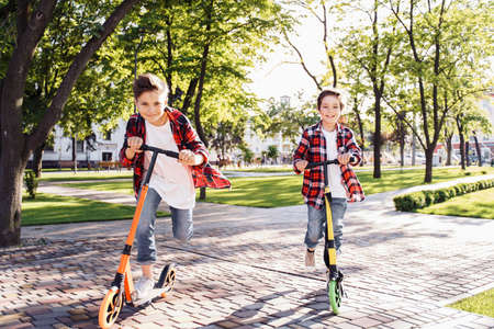 Two happy smiling teenagers brothers, 7 and 8 years old, with speed riding a scooter in the park on a summer evening. Dressed in plaid checkered red shirt, white and jeans. Happy urban childhood.の写真素材