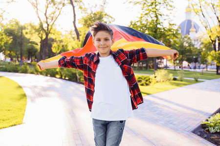 Guy of 9 years old riding skateboard on sunny road in the evening in the city park. Holding fluttering flag of Germany. Happy German day. German language, European education.の写真素材