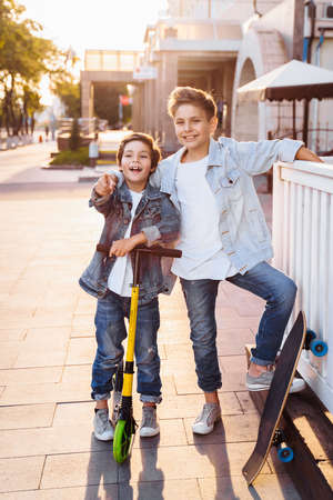 two attractive teenagers of Caucasian nationality 7 and 9 years old standing on sunny evening street in the city. Laughing, pointing fingers forward, holding scooter and skateboard.の写真素材