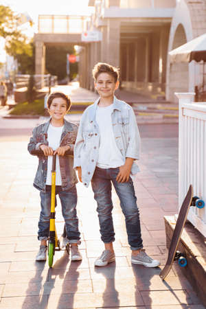 two attractive teenagers of Caucasian nationality 7 and 9 years old standing on sunny evening street in the city. Laughing, pointing fingers forward, holding scooter and skateboard.の写真素材
