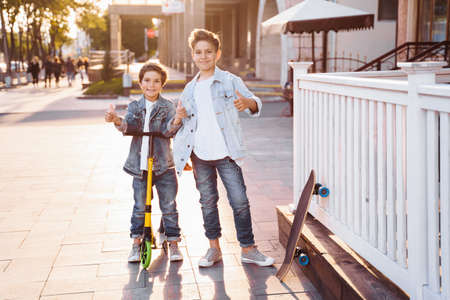 two attractive teenagers of Caucasian nationality 7 and 9 years old standing on sunny evening street in the city. Laughing, pointing fingers forward, holding scooter and skateboard.の写真素材