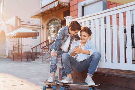 Two attractive caucasian teenagers, wearing denim jackets, pants and white T-shirts sitting near fence, looking to phone. Smiling, laughing, surprising, poking to gadget on evening summer sunset.の写真素材