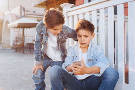 Two attractive caucasian teenagers, wearing denim jackets, pants and white T-shirts sitting near fence, looking to phone. Smiling, laughing, surprising, poking to gadget on evening summer sunset.の写真素材