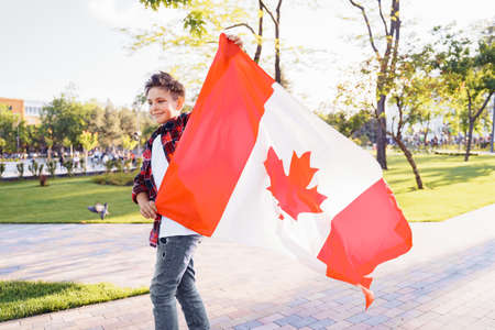 A young guy of 9 years old riding on a sunny road in the evening in the city park, dressed in a flannel plaid shirt. Holding fluttering flag of Canada. Happy Day Independence day to Canada.の写真素材