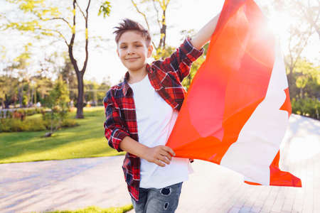 A young guy of 9 years old riding on a sunny road in the evening in the city park, dressed in a flannel plaid shirt. Holding fluttering flag of Canada. Happy Day Independence day to Canada.の写真素材