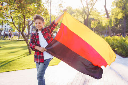 Guy of 9 years old riding skateboard on sunny road in the evening in the city park. Holding fluttering flag of Germany. Happy German day. German language, European education.の写真素材