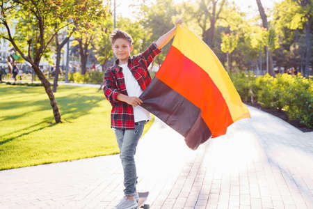 Guy of 9 years old riding skateboard on sunny road in the evening in the city park. Holding fluttering flag of Germany. Happy German day. German language, European education.の写真素材