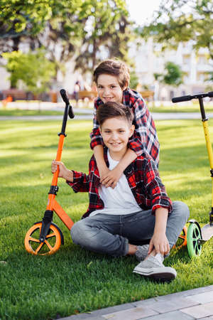 Two european teenagers are sitting on the grass in a summer sunny park. Nearby are their scooters. They laughing, hugging, having fun, spending time on vacation in the city. Active lifestyle concept.の写真素材