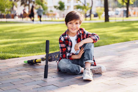 Caucasian teenager sitting on the grass in a summer sunny park with his scooter. Smiling and having fun, spending time on vacation in the city. Active lifestyle concept.の写真素材
