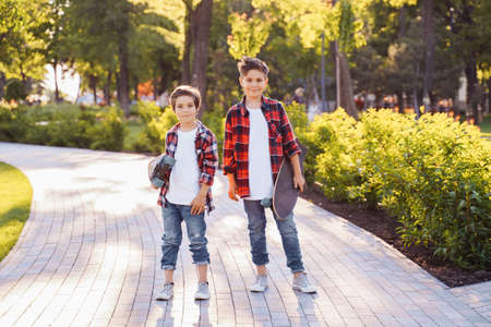 two pretty brothers of Caucasian nationality standing in the park with skateboards in hands. Dressed in jeans and plaid red shirts with white T-shirts. Smiling and enjoying a warm summer evening.の写真素材
