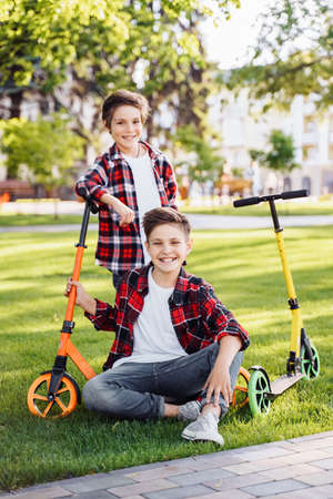Two european teenagers are sitting on the grass in a summer sunny park. Nearby are their scooters. They laughing, hugging, having fun, spending time on vacation in the city. Active lifestyle concept.の写真素材